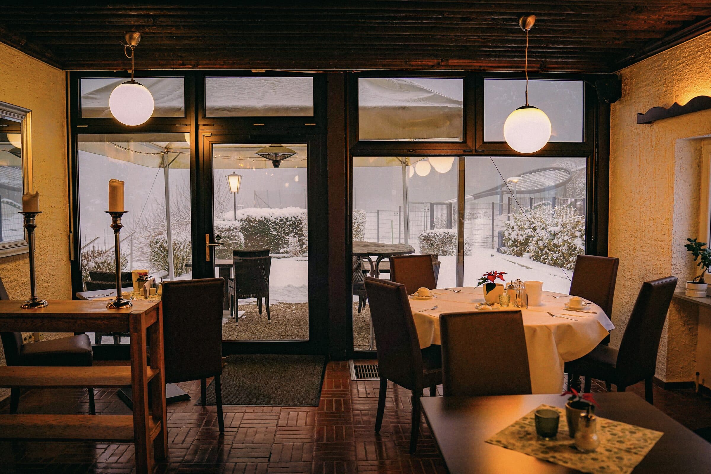 Warm beleuchtete Harzer Stube im Winter: gedeckter Tisch mit Weihnachtsstern, Kerzen und Lichterketten, draußen verschneiter Garten